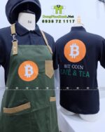 Bộ đồng phục quán cafe gồm áo thun đen có logo Bitcoin và tạp dề màu xanh rêu in logo Bit Coin Cafe & Tea.
