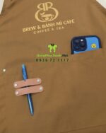 Tạp dề đồng phục màu nâu vàng mộc mạc dành cho nhân viên quán cà phê, in logo Brew & Bánh Mì Cafe.
