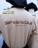 Bộ đồng phục gồm áo thun màu be và tạp dề đen, in logo Sip & Savour nổi bật