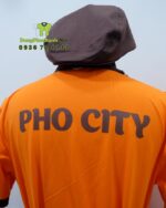 Bộ đồng phục quán ăn Pho City gồm áo thun cam và tạp dề nâu in logo nổi bật