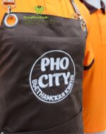 Bộ đồng phục quán ăn Pho City gồm áo thun cam và tạp dề nâu in logo nổi bật